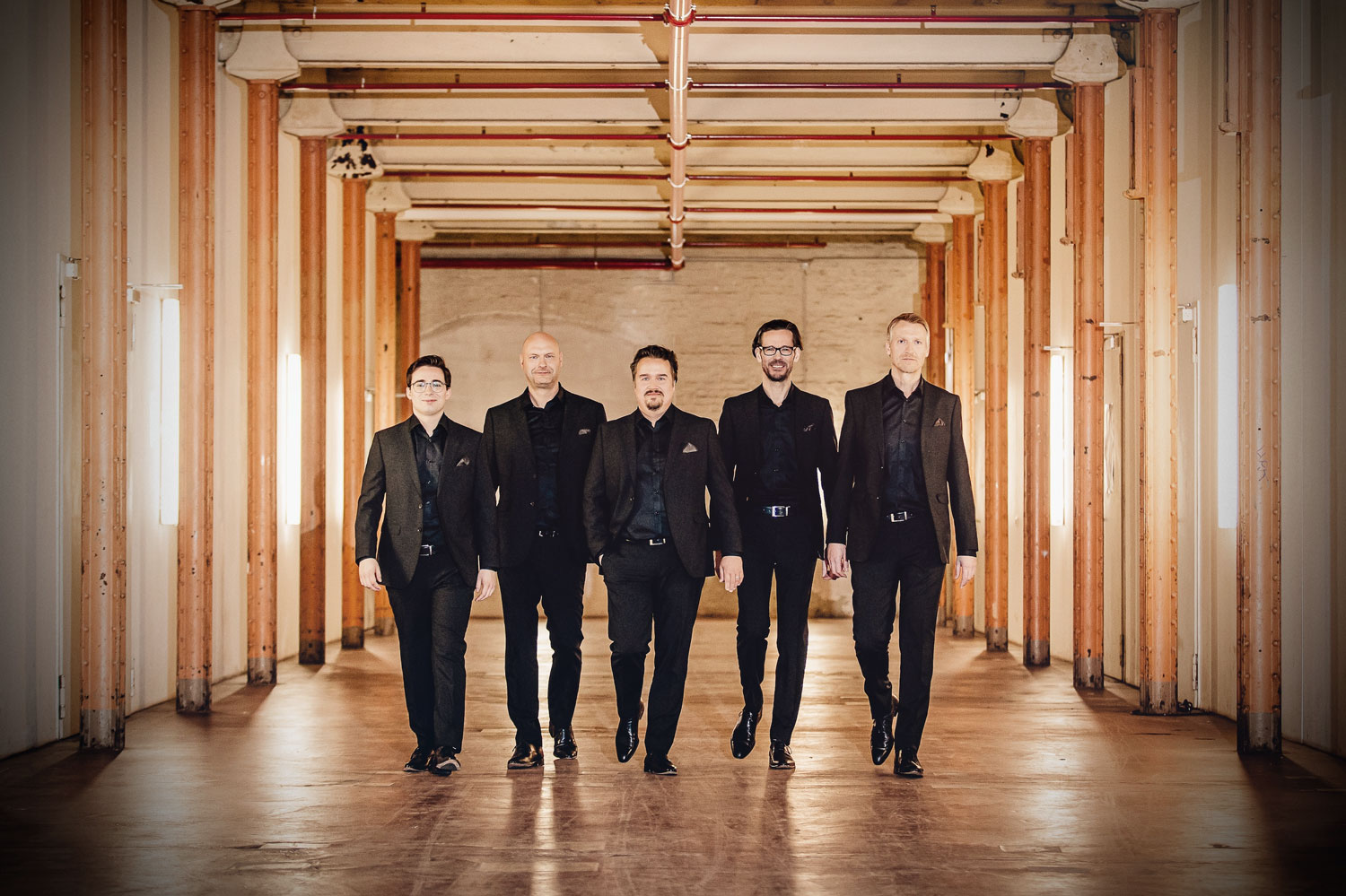 Fünf schwarz gekleidete Herren gehen in einem hell beleuchtetem Gang dem Betrachter entgegen.