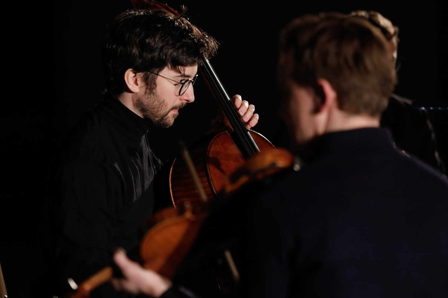 Ein Celisst spielt vor dunklem Hintergrund vor ihm ist ein Geger von hinten zu sehen