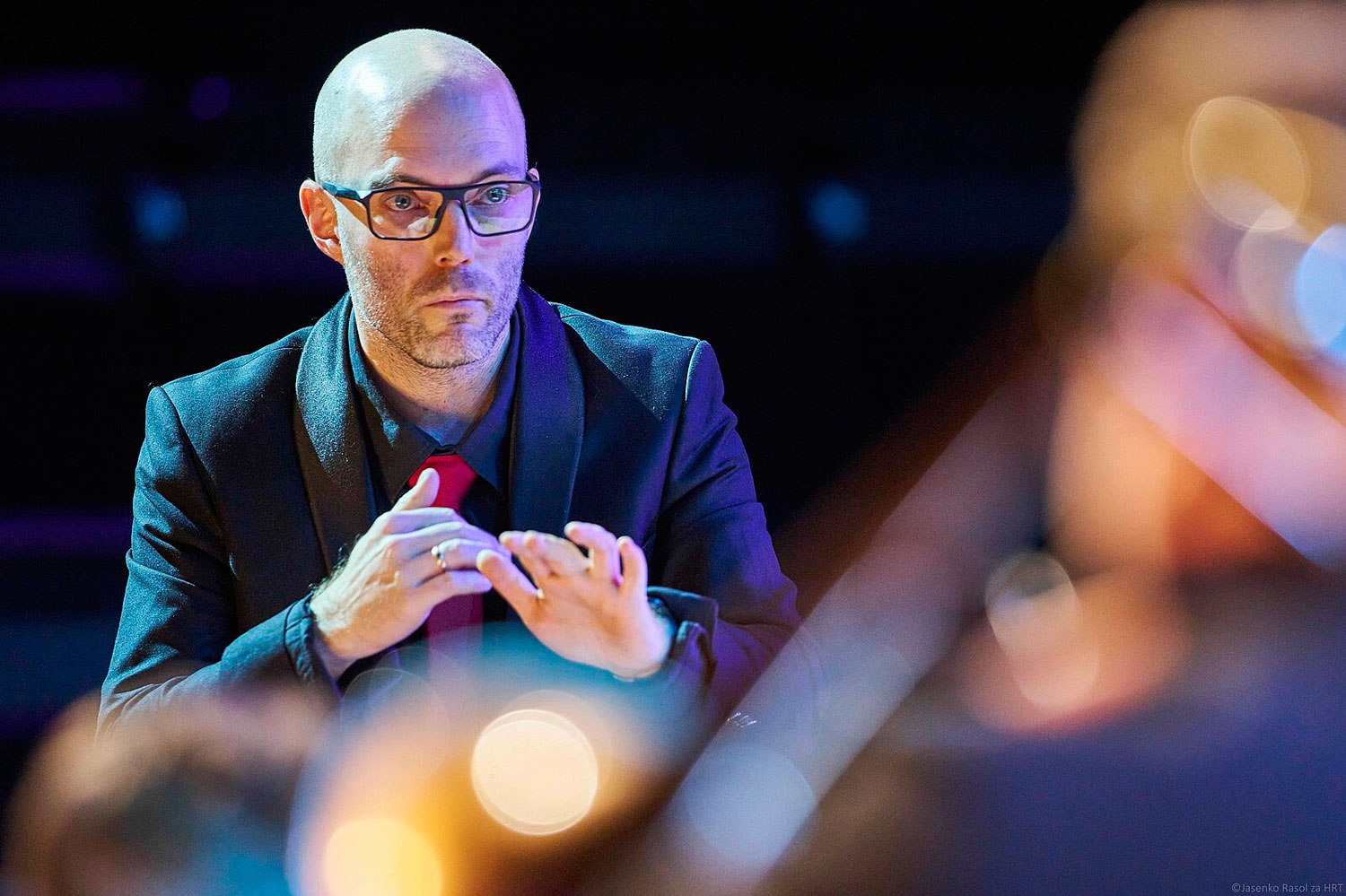 Ein Mann mit Brille dirigiert mit konzerntriertem Blick. Im Vorderdrund sind unschafre Andeutungen von Musikinstrumenten zu erkennen, deren Reflektionen dem Foto ein schönes Bokeh verleihen.