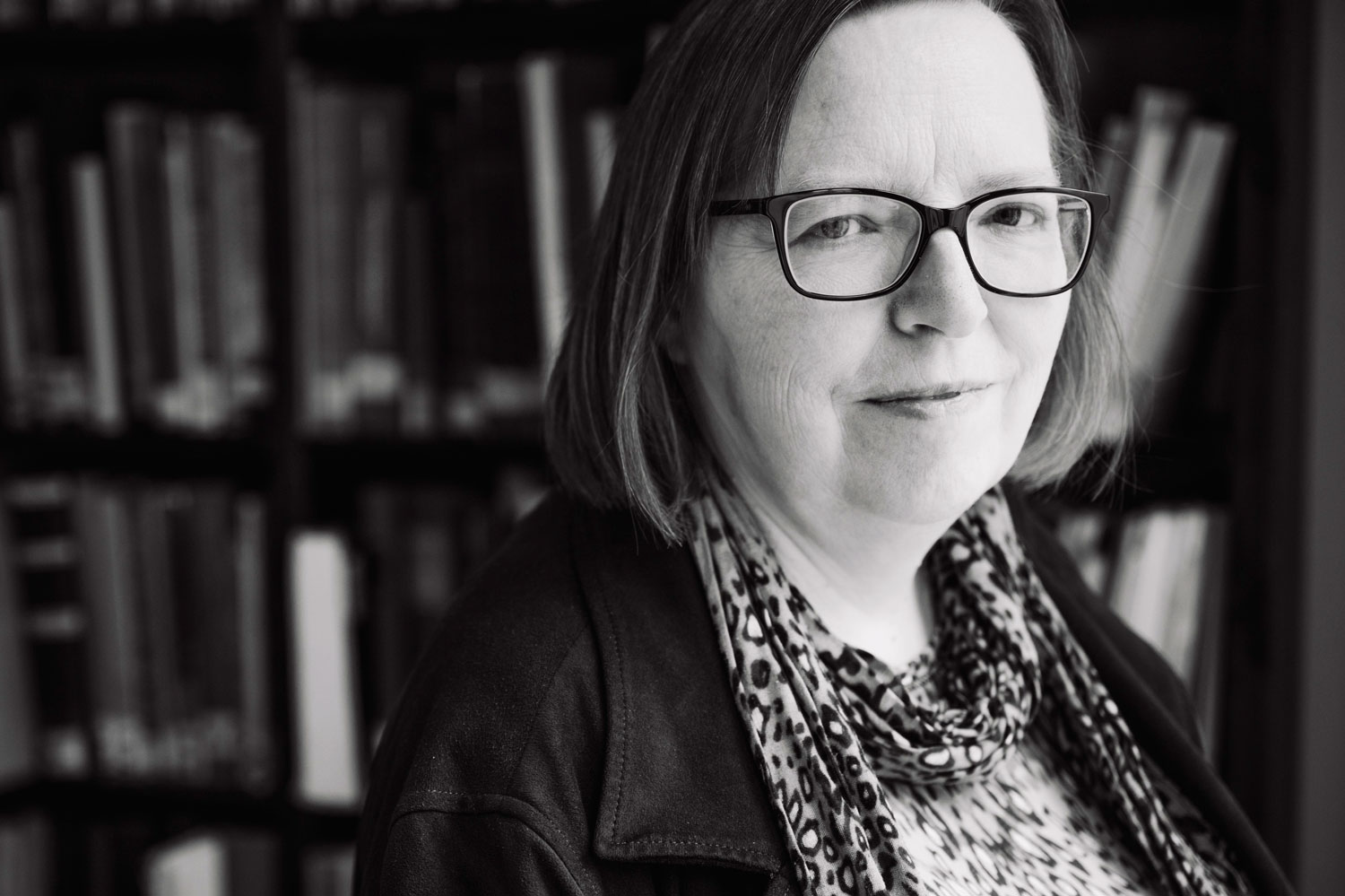 Portraitfoto in schwarzweiß von Christine Siegert, die vor einer Bücherwand stehend in die Kamera blickt.