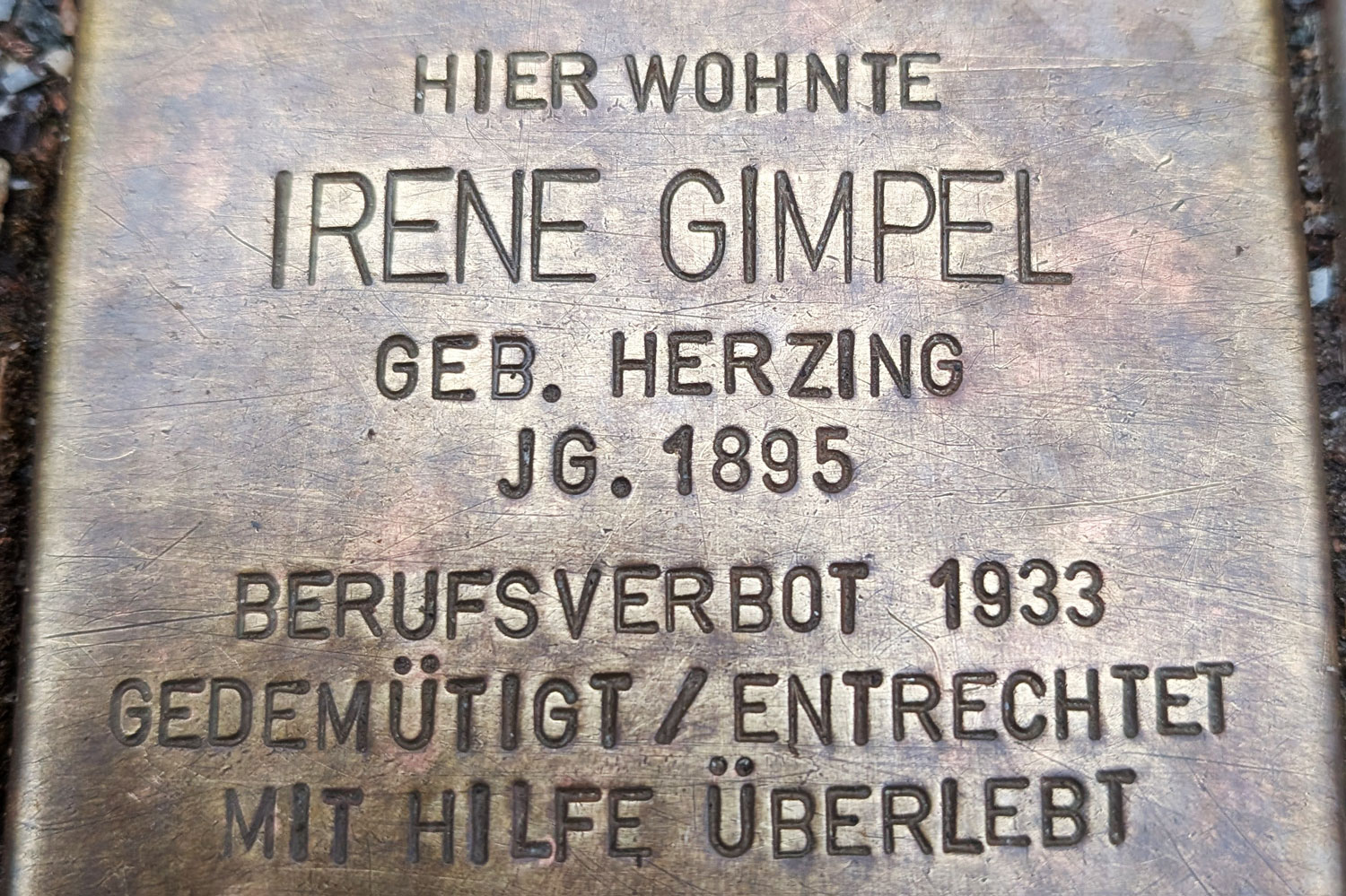 Zu sehen ist ein messingfarbener Stolperstein mit der Inschrift: Hier wohnte Irene Gimpel, geb. Herzing, JG. 1895, Berufsverbot 1933, Gedemütigt / entrechtet, mit Hilfe überlebt