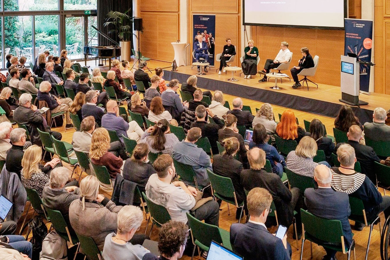 Blick von erhöhter Position in einen Konferenzraum. Im Hintergrund sind fünf Personen auf einem Podium sitzend zu erkennen. Davor ein mit Publikum gut gefüllter Saal